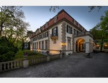 Historisches Gebäude mit heller Fassade, roten Dachziegeln und großen Fenstern, umgeben von einem Garten mit Bäumen. Im Eingangsbereich befindet sich ein Arkadengang mit Laternen, und eine Balustrade grenzt den Vorplatz ab.