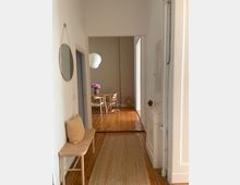 Heller Flur mit Holzboden und einem geflochtenen Teppich, links steht eine Sitzbank mit einem Kissen, rechts ein schmaler Einbauschrank. Im Hintergrund ein angrenzender Raum mit einem Holztisch, Holzstühlen und einer weißen Hängelampe.