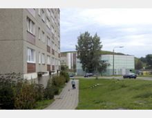 Wohnsiedlung mit einem mehrstöckigen Plattenbau auf der linken Seite, einem Gehweg, auf dem eine Person mit einem kleinen Hund läuft, sowie flachen, grünen Rasenflächen und einem modernen, hellgrünen Gebäude im Hintergrund. Einige geparkte Autos und ein Baum sind ebenfalls zu sehen.