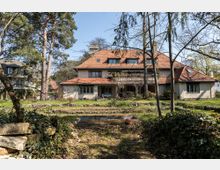 Großes, zweigeschossiges Wohnhaus mit rotem Ziegeldach und mehreren Gauben, umgeben von einem großzügigen Garten mit Bäumen, Sträuchern und einer kleinen Teichanlage im Vordergrund. Das Gebäude befindet sich in einer ruhigen, grünen Wohngegend.