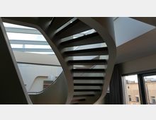 Moderne Innenansicht mit einer freitragenden Wendeltreppe aus dunklem Holz und einem geschwungenen Metallgeländer. Im Hintergrund sind eine Glasdecke, große Fenster sowie ein Teil der Stadtansicht sichtbar.