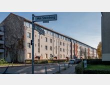 Mehrstöckiges Wohngebäude in einer ruhigen Straßenumgebung, mit teils verputzter und teils rot verklinkerter Fassade. Im Vordergrund Straßenschilder, vereinzelte Autos und Gehwege, umgeben von kleinen Grünflächen und Büschen.