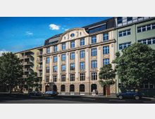 Mehrstöckiges städtisches Gebäude mit klassischer Sandsteinfassade, symmetrischen Fensterreihen und rundbogigen Öffnungen im Erdgeschoss, darüber ein Dachaufbau mit Gauben. Daneben angrenzen moderne Wohngebäude mit Balkonen; davor verläuft eine breite Straße mit parkenden Autos und Bäumen am Gehweg.