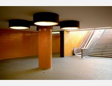 U-Bahn-Unterführung mit orangefarbenen, gefliesten Wänden, einer zentralen runden Säule mit drei großen Leuchten und einer Treppe sowie einer Rolltreppe im Hintergrund. Ein Schild mit der Aufschrift „Aufzug“ ist sichtbar.