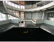 Innenansicht eines modernen Gebäudes mit einer offenen, runden Bauweise. Zu sehen sind zwei übereinanderliegende Etagen mit Metallgeländern, eine geschwungene Treppe links, große Fenster mit Lamellen und eine schwarze, erhöhte Plattform im Vordergrund.