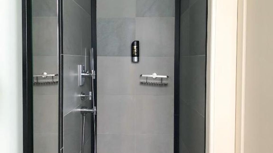 Schmale, moderne Dusche mit grauen Wand- und Bodenfliesen, schwarzer Randgestaltung und bodengleichem Ablauf. Links befindet sich eine Glasabtrennung mit Chromarmaturen und Handbrause, oben ein fest montierter Duschkopf.