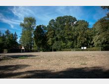 Weitläufiger, trockener Platz mit spärlichem Grasbewuchs, umgeben von hohen Bäumen. Im Hintergrund sind ein kleiner gemauerter Pavillon, Hinweistafeln und eine Bank zu sehen.