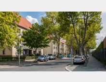 Straßenszene in einem Wohngebiet mit mehrstöckigen, hellen Wohngebäuden und roten Ziegeldächern, flankiert von großen Bäumen. Am Straßenrand stehen parkende Autos, und ein Gehweg verläuft entlang der rechten Seite.