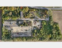 Luftaufnahme eines verlassenen Industriegeländes mit mehreren rechteckigen Gebäuden und großen betonierten Flächen, umgeben von dichtem Grün und Bäumen. Im oberen Bildbereich verläuft eine Straße, rechts grenzt das Gelände an ein landwirtschaftliches Feld.