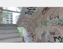 Verlassene Treppe eines baufälligen Gebäudes mit an den Wänden und der Decke angebrachten Graffitis. Im Hintergrund sind weitere verfallene Gebäude sowie Bäume und Vegetation sichtbar.
