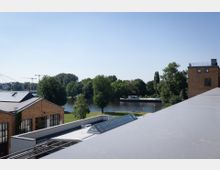 Ansicht eines Flussufers mit Bäumen und einer Anlegestelle, umgeben von modernen Gebäuden mit Backsteinfassaden und Glaselementen im Vordergrund. Der Himmel ist klar, und im Hintergrund sind Kräne und Wohngebäude zu erkennen.