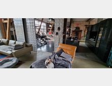 Wohnbereich in einem loftartigen Gebäude mit offenen Räumen und industriellem Design. Im Vordergrund steht ein beiger Sessel mit einer Decke und einem kleinen Hund darauf, daneben ein Sofa, im Hintergrund sind eine Treppe, große gläserne Trennwände, eine Küche mit schwarzen Schränken und eine Ziegelwand mit antiken Möbeln zu sehen.