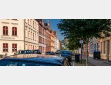 Straßenszene mit historischen Gebäuden in Wittenberge; die Hausfassaden sind in hellen Farben mit roten Fenstersprossen gestaltet. Auf der Straße parken Autos, während an der rechten Seite Bäume und Mülltonnen stehen.