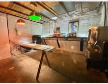 Barbereich in einem industriellen Raum mit unverputzten weißen Wänden, einer einfachen Holzdecke und einem Betonboden. Im Vordergrund ein langer Holztisch auf Böcken, im Hintergrund eine Bar mit Getränkeflaschen, einer Spüle aus Metall und bunter Beleuchtung.