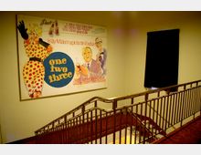 Treppenhaus mit rotem Teppichboden und Metallgeländer, an der Wand ein großformatiges Poster des Films „One, Two, Three“ mit farbenfroher Illustration und Schriftzug; daneben ein kleines, verdunkeltes Fenster.