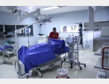 Krankenhaus-Operationssaal mit einem mit blauem Tuch abgedeckten Operationstisch, medizinischen Geräten wie Monitoren und Infusionsständern sowie einem Arzt, der im Hintergrund arbeitet. Die Wände sind mit hellblauen Kacheln verkleidet, und medizinische Ausrüstung steht auf Regalen und Wagen bereit.