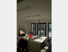 Ein kleiner, heller Büroraum mit mehreren zusammengestellten Tischen, auf denen Büromaterialien und Laptops verteilt sind. Große Glasfenster mit Türen führen zu einem kleinen Balkon, und an der Decke sind rechteckige Leuchten angebracht.