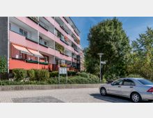 Mehrstöckiges Wohngebäude mit rosa Fassaden und Balkonen, teils mit Markisen und Pflanzen dekoriert. Im Vordergrund ein gepflasterter Bürgersteig, ein parkendes Auto sowie grüne Sträucher und Bäume.