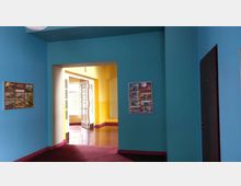 Ein Innenraum mit blau gestrichenen Wänden, einem roten Teppichboden und gelben Wänden im angrenzenden Bereich. Eine weiße Tür mit Glasfenstern führt in einen hellen Raum, an den Wänden hängen Informationsposter.