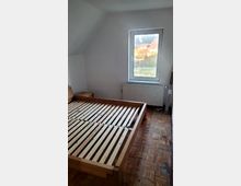 Ein einfach eingerichtetes Schlafzimmer mit einem Holzlattenrost-Bettgestell auf einem Parkettboden. Ein Fenster mit Blick auf eine ländliche Umgebung befindet sich oberhalb eines weißen Heizkörpers.