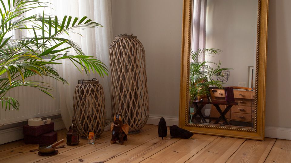 Helle Zimmerecke mit breitem Holzdielenboden, bodentiefem Fenster mit transparenten Vorhängen und einer großen Zimmerpflanze. Daneben stehen zwei geflochtene, hohe Lampenschirme oder Körbe und ein großer, goldgerahmter Standspiegel, der eine Kommode und weitere Pflanzen im Raum reflektiert.