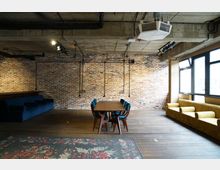 Ein modern gestalteter Raum mit sichtbarer Ziegelwand, Betondecke und freiliegenden Rohren. In der Mitte steht ein Holztisch mit blauen Stühlen, links und rechts befinden sich Sitzbänke in Blau und Gelb, während Fenster rechts viel Tageslicht hereinlassen.