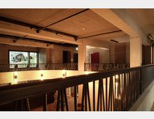 Obergeschoss eines modernen Gebäudes mit industriellen Designelementen wie freiliegenden Betonwänden und einer schwarzen Metallbalustrade. Im Hintergrund sind ein großes Fenster mit einem Fischmotiv und Türen in dunklen Farbtönen sichtbar.