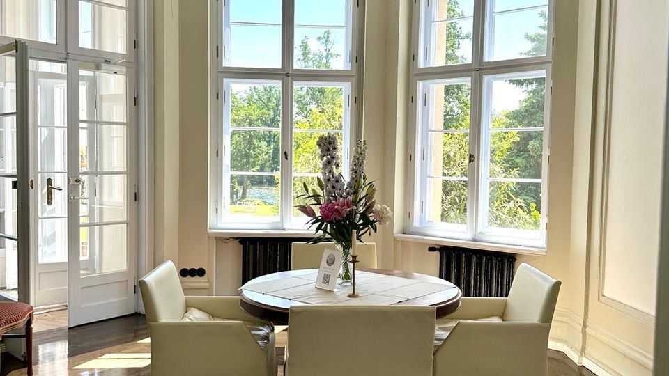 Heller Speiseraum in einem Altbau mit hohen Decken, Stuck und zentraler Hängelampe sowie großen Sprossenfenstern in einem Erker. In der Mitte steht ein runder Tisch mit vier Stühlen auf einem Holzparkettboden; seitlich führt eine Glastür nach außen.