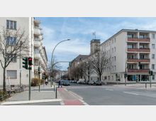 Straßenszene in einer Stadt mit Wohnhäusern und Bäumen, im Hintergrund ein Gebäude mit Uhrturm und gehisster Fahne. Zu sehen sind eine Kreuzung mit Fußgängerampeln, geparkte Autos und vereinzelt Passanten.