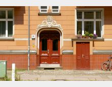 Fassade eines Wohnhauses mit orangefarbenem Putz und roten Kacheln im unteren Bereich. Zentrales Element ist eine reich verzierte Holzeingangstür mit geschwungener weißer Stuckverzierung, daneben ein Fenster mit Blumenkästen sowie eine kleinere rote Tür, vor der ein Fahrrad angelehnt ist.