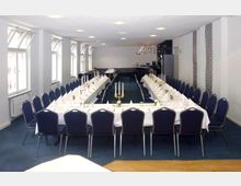 Ein länglicher Veranstaltungsraum mit zwei langen, festlich gedeckten Tischen, die in U-Form angeordnet sind, umgeben von blauen Polsterstühlen. Im Hintergrund befindet sich eine Bar, und an der linken Seite des Raums sind mehrere Fensterreihen sichtbar.