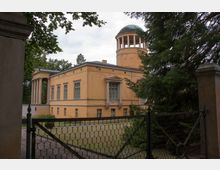 Gelbes, zweistöckiges klassizistisches Gebäude mit Säulenportikus und einer Kuppel auf einem runden Turm, umgeben von Bäumen und einer schmiedeeisernen Gittertor-Einfriedung. Im Vordergrund ein kleiner Garten mit Rasen.