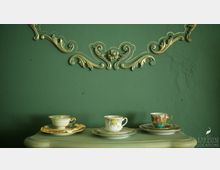 Drei verzierte Teetassen mit Untertassen stehen auf einem grünen Holztisch vor einer dunkelgrünen Wand mit goldenen, floralen Stuckverzierungen.