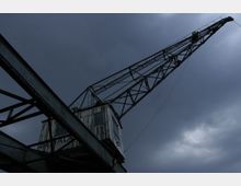 Ein alter, rostiger Hafenkran mit einer langen Schwenkarme aus Metallunterkonstruktionen vor einem bewölkten, düsteren Himmel.