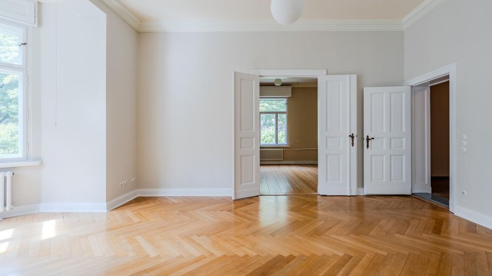 Heller, leerer Altbau-Wohnraum mit hohen Decken, Stuckleisten und Fischgrätparkett. Links ein großes Fenster, mittig eine doppelflügelige Kassettentür zu einem weiteren Raum mit Fenster und Heizkörper.