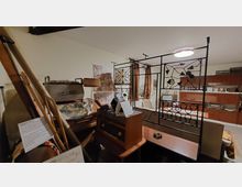 Wohnzimmer im Retro-Stil mit alten Möbeln und Einrichtungsgegenständen aus der Mitte des 20. Jahrhunderts. Im Vordergrund stehen ein altes Radio und ein Bügeleisen auf einem Holzschrank, während der Raum durch eine dekorative Metalltrennwand mit floralen Mustern geteilt wird. Im Hintergrund sind eine Schrankwand, ein Fernseher und weitere Vintage-Elemente zu sehen.