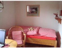 Kleines Schlafzimmer mit einem Holzbett und rot-weiß kariertem Bettzeug. Im Vordergrund ein geflochtener Stuhl mit passendem Kissen, ein runder Holztisch und ein Laptop, an der Wand ein Regal mit Deko und eine gerahmte Fotografie.