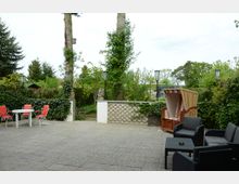 Gepflasterte Terrasse mit Gartenmöbeln, darunter ein Strandkorb, ein schwarzes Sitzmöbel-Set und ein Tisch mit roten Stühlen. Die Umgebung ist von Pflanzen, einer Hecke und kleinen Bäumen umgeben, mit lamellenartigen Wänden und Lampen an den Rändern.