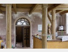 Innenraum eines historischen Gebäudes mit verwitterten Wänden und Holzbalkendecke. Zu sehen sind eine Arkadentür mit Rundbogen und Glaseinsatz, Stützbalken mit geschnitzten Details sowie ein Tresenbereich mit einfachen Einrichtungsgegenständen und einem Wasserkocher.