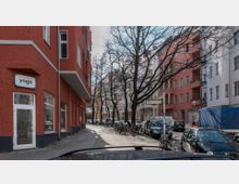 Straßenszene in einer Wohngegend mit Mehrfamilienhäusern in roten, rosa und weißen Fassaden. Auf dem Gehweg stehen Fahrräder und Bäume ohne Blätter, entlang der Straße sind geparkte Autos zu sehen; ein Schild mit der Aufschrift „Großgörschenstraße“ markiert die Ecke.