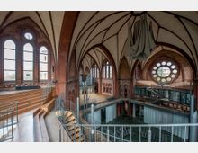 Innenraum einer umgebauten Kirche mit neugotischer Architektur, gekennzeichnet durch spitzbogige Fenster, rotes Ziegelmauerwerk und ein großes Rundfenster. Moderne Elemente wie Metalltreppen, Geländer und ein abgetrennter Bereich mit Stühlen ergänzen die historische Struktur.