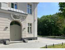 Teilansicht eines historischen Gebäudes mit heller Fassade, großen Fenstern und einem verzierten Eingangsbogen mit dekorativer Beschriftung über der Tür. Rechts neben dem Gebäude ist eine gepflasterte Fläche und dahinter eine Baumreihe zu sehen.
