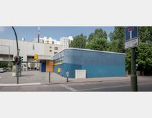 Straßenszene mit einem blauen, kubusförmigen Gebäude mit gelben Elementen und moderner Architektur. Im Hintergrund sind Wohngebäude und Bäume sichtbar, vorne ein Fußgängerüberweg mit Ampel und ein Verkehrszeichen.