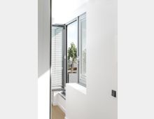 Heller Innenraum mit weiß gestrichenen Wänden und Holzboden, ein geöffnetes Fenster mit grauem Rahmen und Jalousien zeigt einen Blick auf eine Terrasse mit Pflanzen.