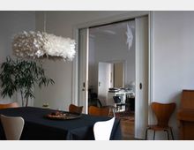 Esszimmer mit einem schwarzen Tisch und Holzstühlen, einer weißen Federlampe an der Decke und einer Topfpflanze in der Ecke. Im Hintergrund ist durch eine offene Schiebetür ein weiteres Zimmer mit Büchern und modernen Möbeln sichtbar.