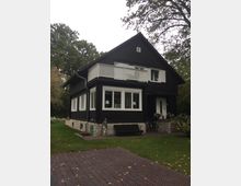 Schwarzes zweistöckiges Holzhaus mit weißer Fenster- und Balkonverkleidung, auf einem Fundament aus Stein. Vor dem Haus befinden sich eine Rasenfläche, eine Steinbank und eine Statue, umgeben von hohen Bäumen.