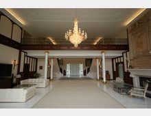 Großer, eleganter Eingangsbereich mit Marmorboden, einer breiten Teppichmatte und weiß-goldenen Säulen, die eine Galerie mit verzierten Geländern tragen. Ein großer Kronleuchter hängt von der Decke, und die Einrichtung umfasst helle Sofas, einen Kamin und klassische Möbel.