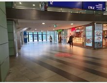 Bahnhofspassage mit Geschäften und Glas-Elementen, darunter eine beleuchtete Bäckerei mit Auslage und Werbeschilder. Im Hintergrund sind automatische Glastüren und der Ausgang Richtung Friedrichstraße sichtbar.