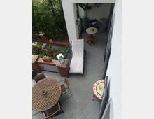 Ein Balkon mit grauem Bodenbelag, einer weißen Wand und mehreren Sitzmöglichkeiten. Es gibt einen runden Holztisch mit Stühlen, eine Liege, kleine Mosaiktische und Blumen sowie eine gemauerte Brüstung aus roten Ziegelsteinen mit Pflanzen darauf. Im Hintergrund befindet sich eine überdachte Nische mit Dekoration und einem Abfallbehälter.