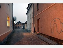 Schmale, gepflasterte Gasse mit einfachen, verputzten Wohnhäusern auf beiden Seiten, graffitibeschmierter Wand rechts und einem beleuchteten Fenster links; im Hintergrund ein älteres Haus mit Satteldach.